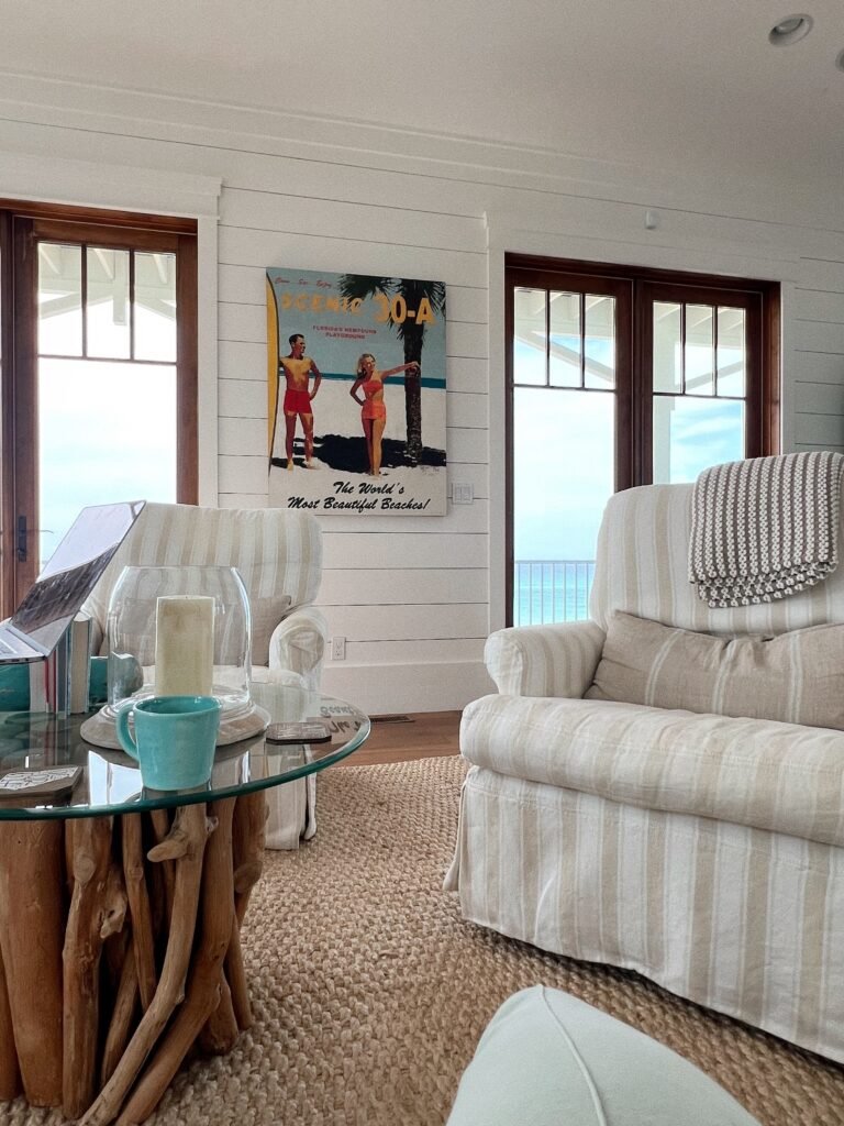 Coastal living room with cream linen slipcovered armchairs, a driftwood-base glass side table, jute rug, and vintage 30A beach art framing an ocean view.